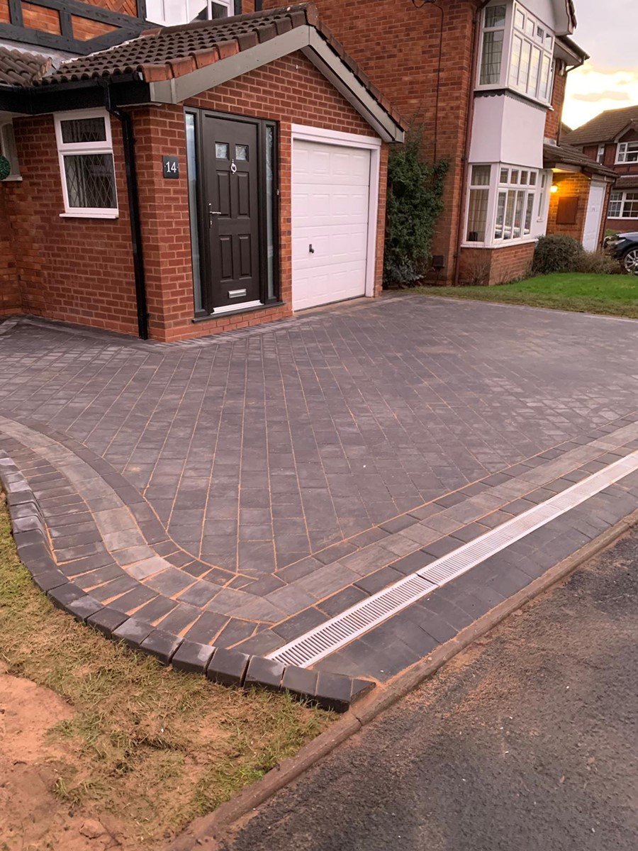 Charcoal block paved driveway with curved edges and a drainage channel at the road